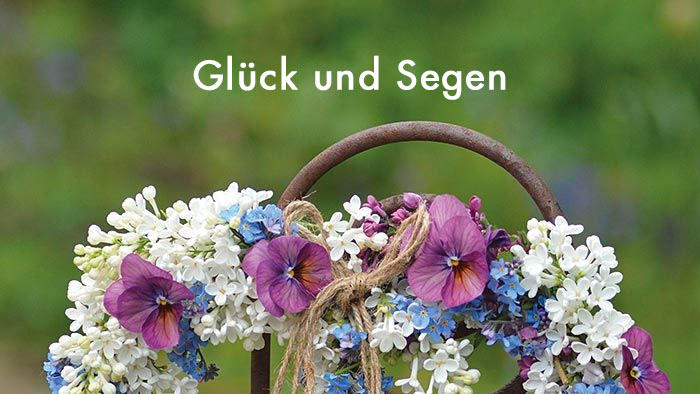 weiße Schrift Glück und Segen - im Hintergrund eine Blumengirlande um einen rostigen Metallstab