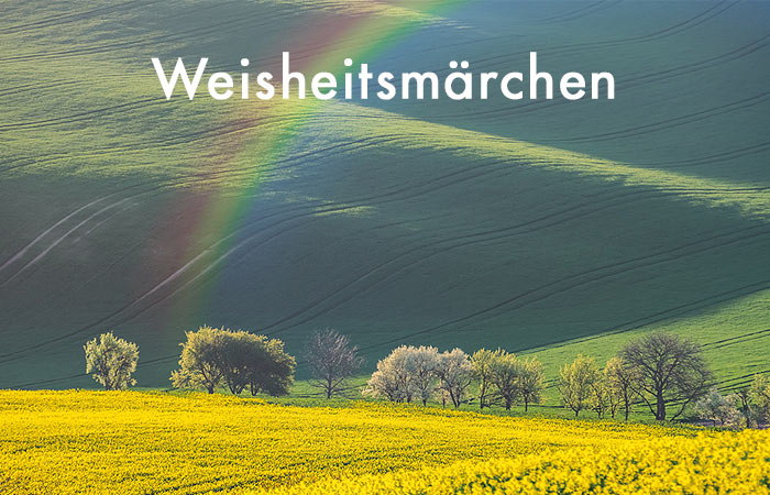 weiße Schrift Weisheitsmärchen - im Hintergrund gelb blühendes Rapsfeld vor grünen Hügeln und einem Regenbogen