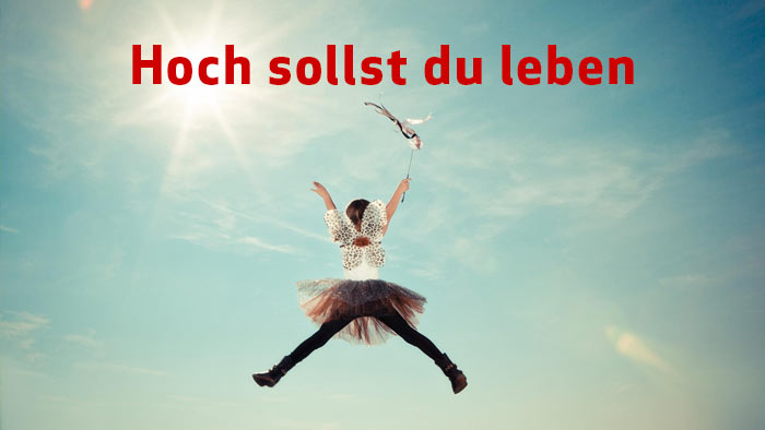 rote Schrift Hoch sollst du leben - im Hintergrund Kind mit Kostümflügeln, das in die Luft springt